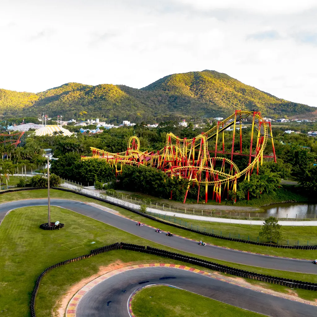 A yellow roller coaster with red supports stands behind a go-kart track, with wooded hills beyond.