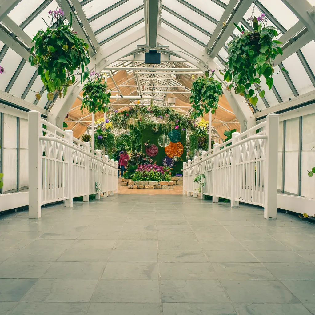 A glass-roofed garden walkway leads past hanging plants to a flower display.