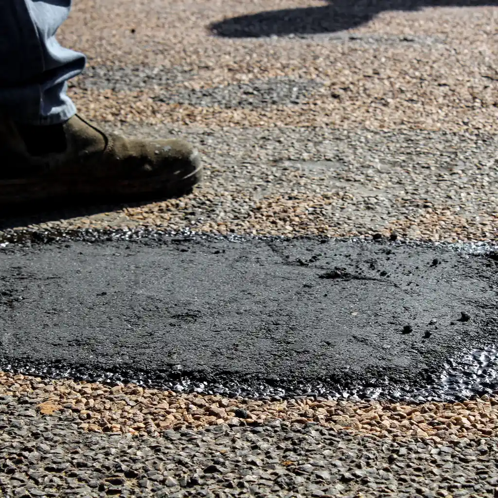 Fresh dark asphalt patches fill localized road defects beside a worker’s boots.