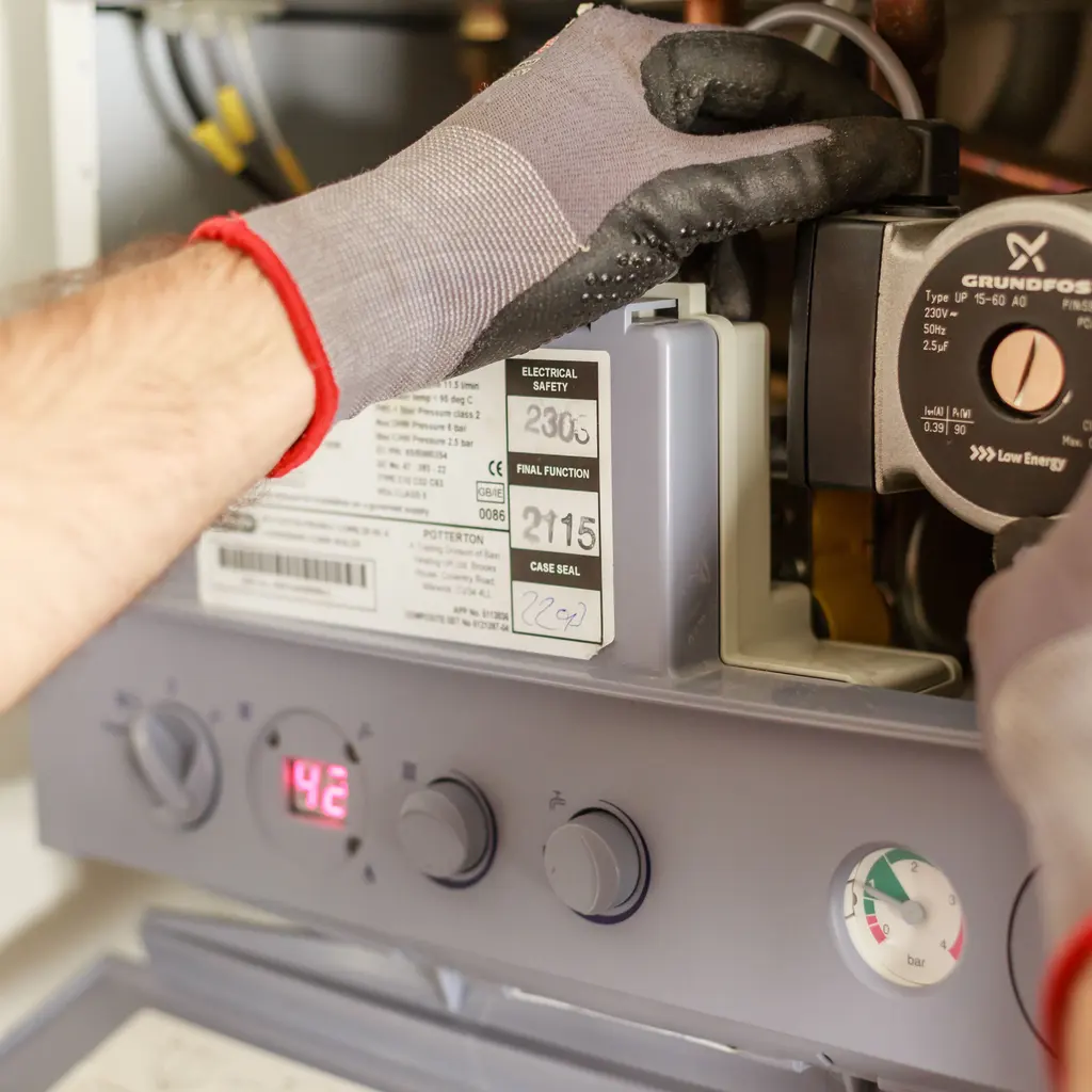 Gloved hands adjust a heating boiler’s pump and digital control assembly during maintenance.