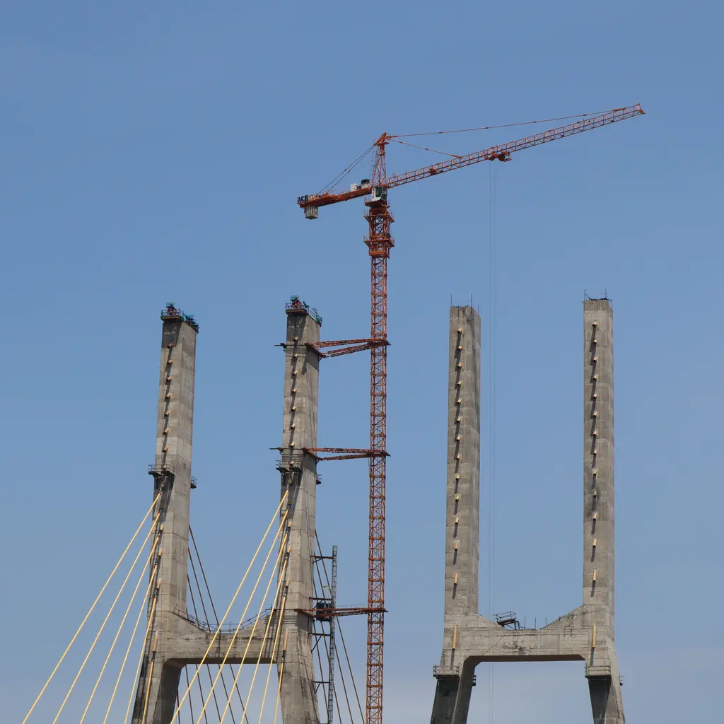 Construction crane works beside unfinished cable-stayed bridge pylons under a blue sky.