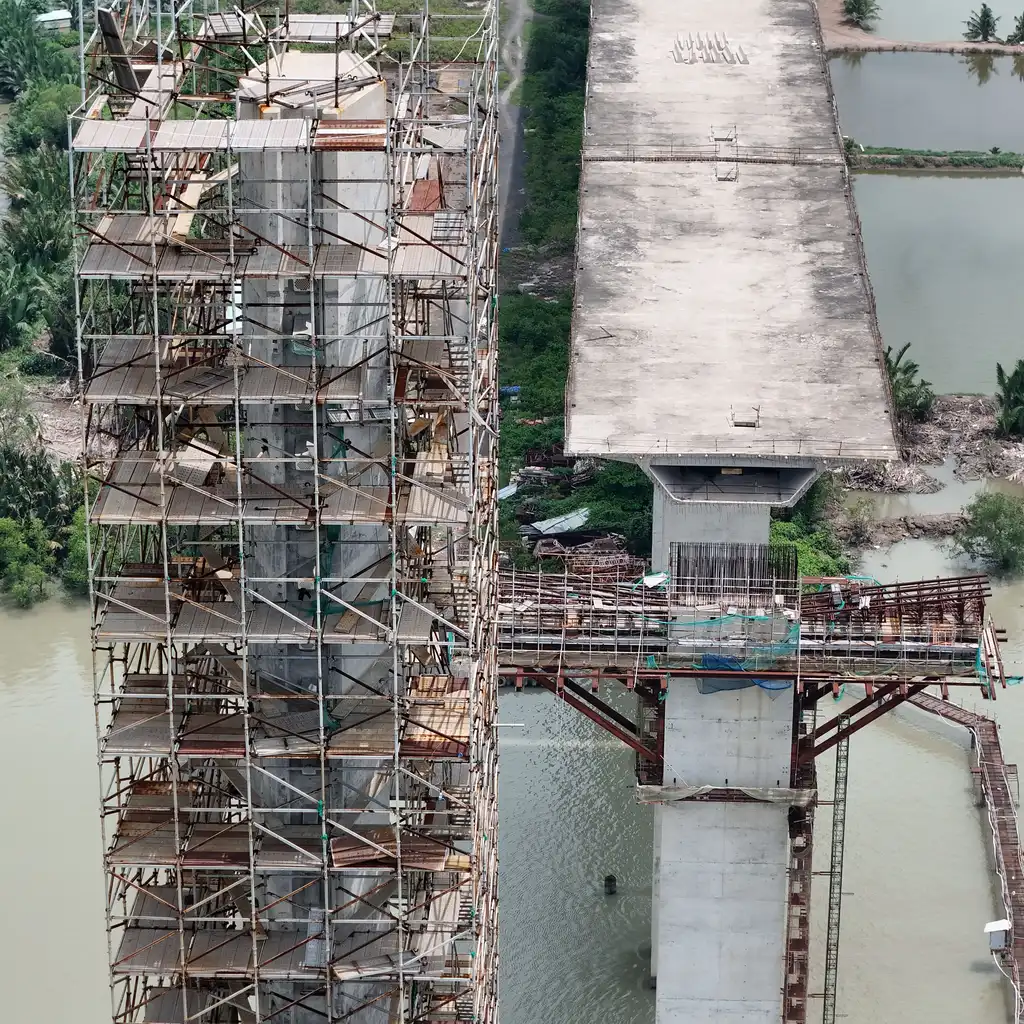 Overhead view shows scaffolded bridge piers and an unfinished concrete deck over a river.