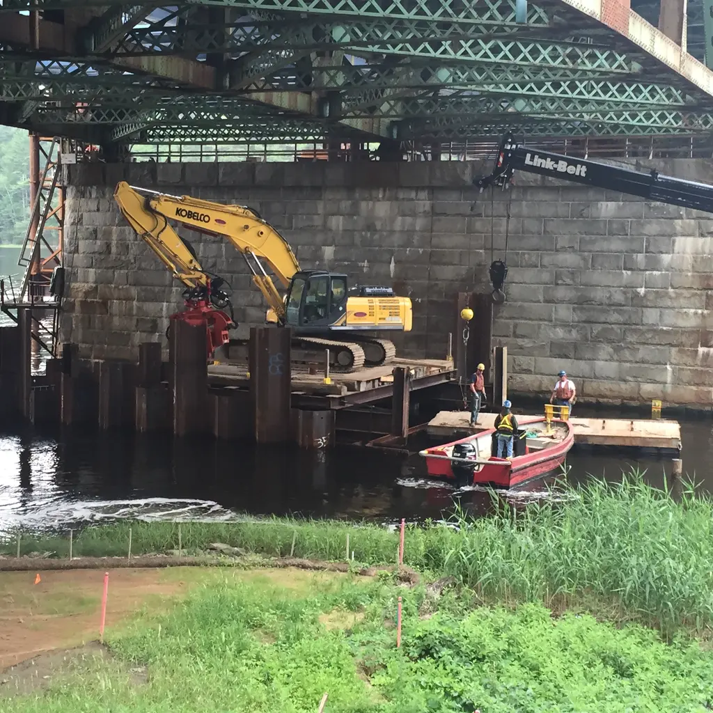 Steel cofferdam walls and an excavator surround in-water bridge-pier construction beneath an existing bridge.