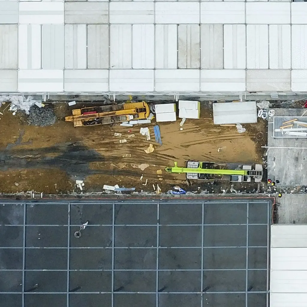 Aerial view of cranes and staged materials between unfinished roofs at a large building site.