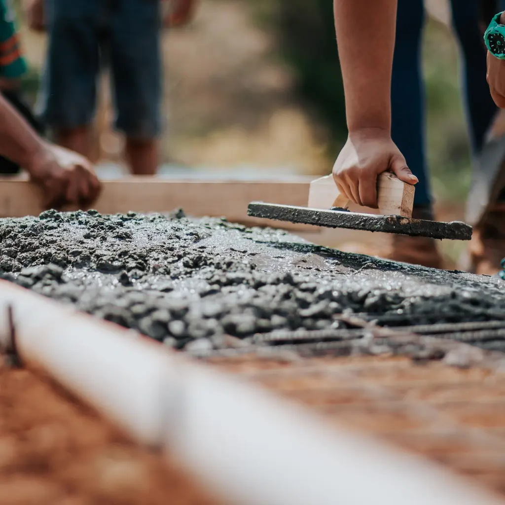 Hands use a concrete float and timber screed to level freshly placed concrete inside an edge form.