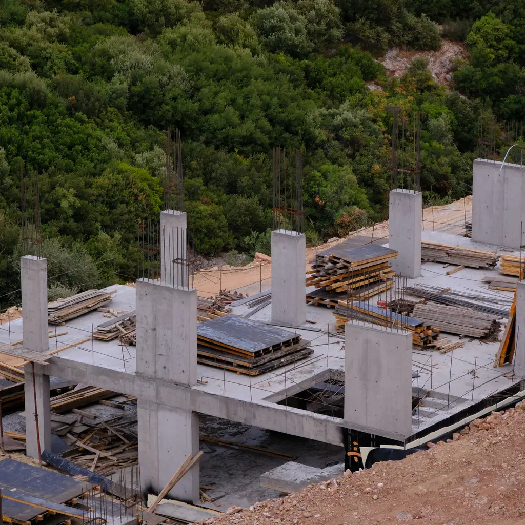Concrete foundation slabs and columns are being built beside excavation in a forested site.