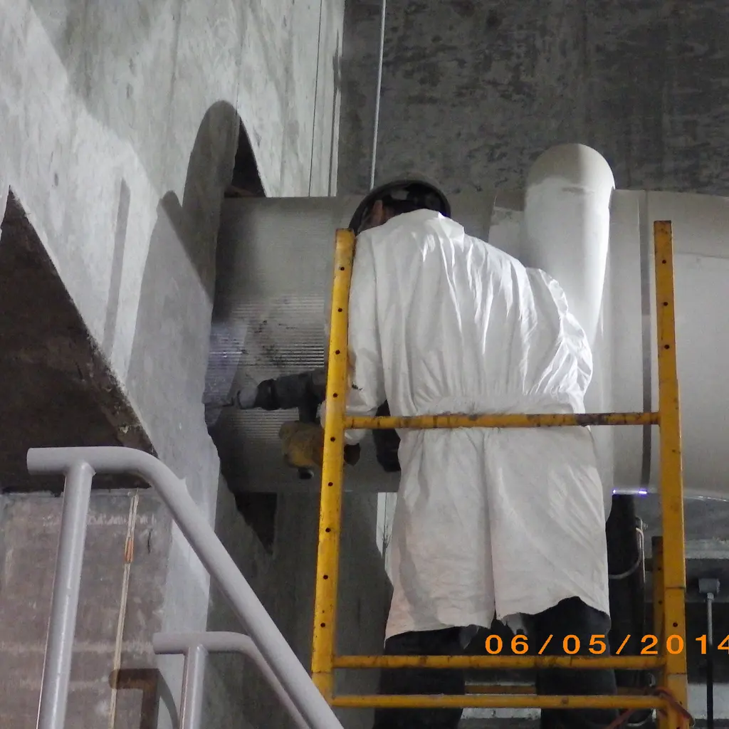 A protected worker carries out concrete repair around a large wall pipe opening from a small scaffold.