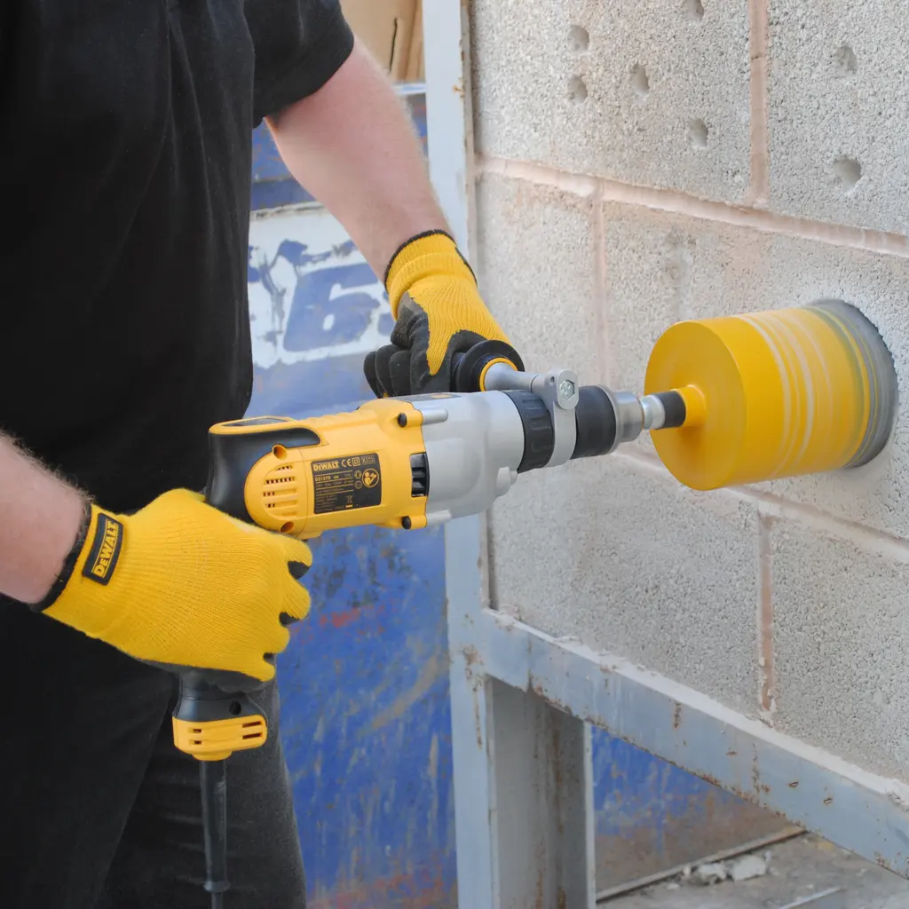 Gloved hands operate a diamond core drill against a concrete-block wall with a large circular drilling barrel.