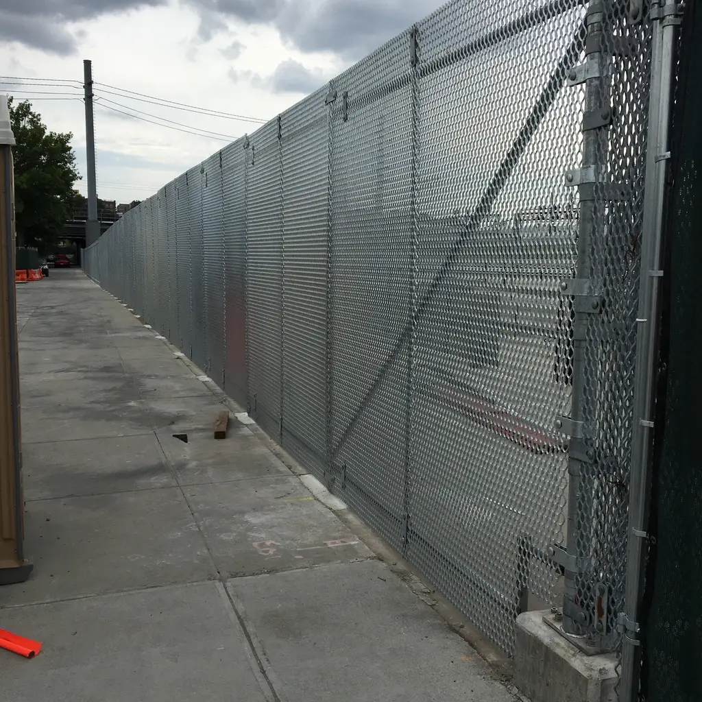 A tall chain-link security fence with privacy inserts and diagonal bracing along a sidewalk.
