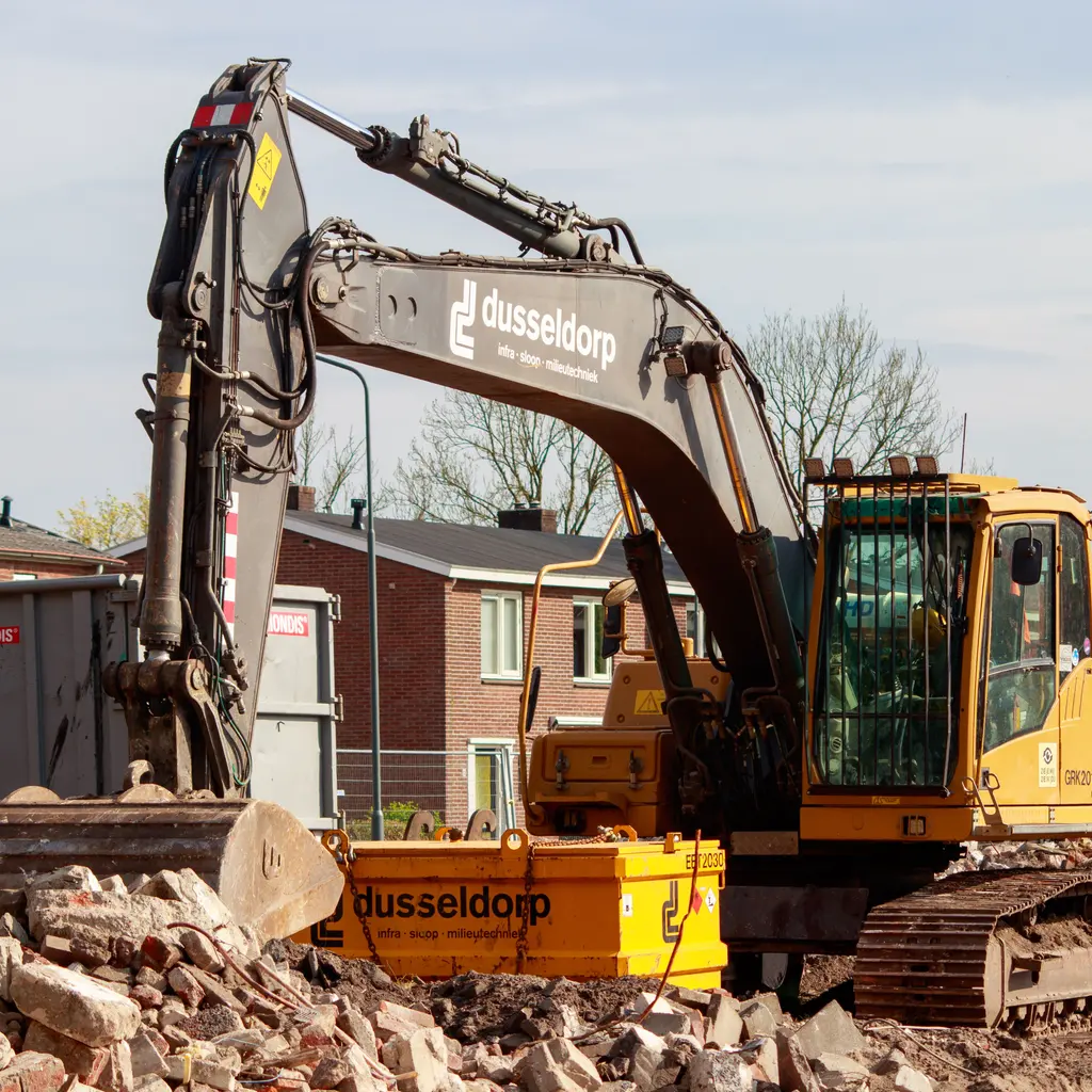 Tracked excavator and bucket sit among concrete rubble during urban construction-site clearing.