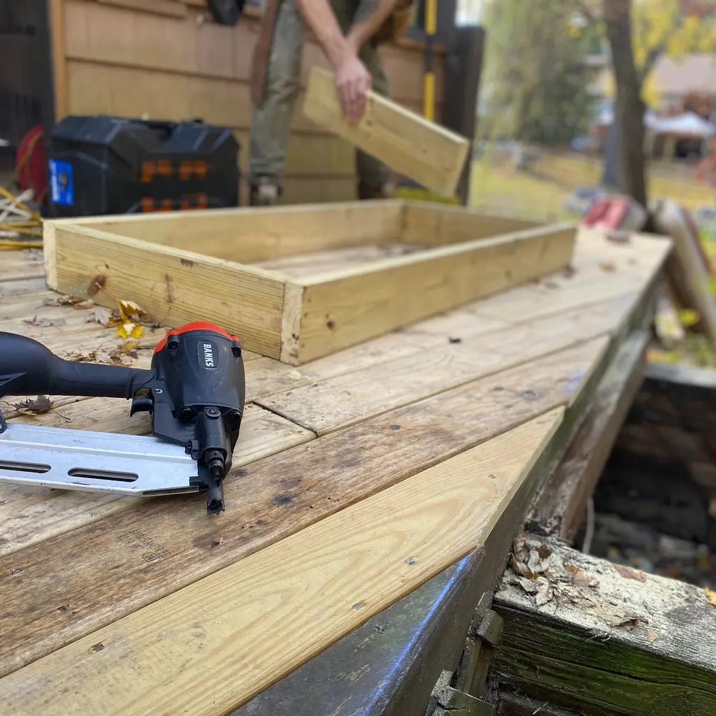 Pneumatic nail gun rests on timber decking beside a deck frame under construction.