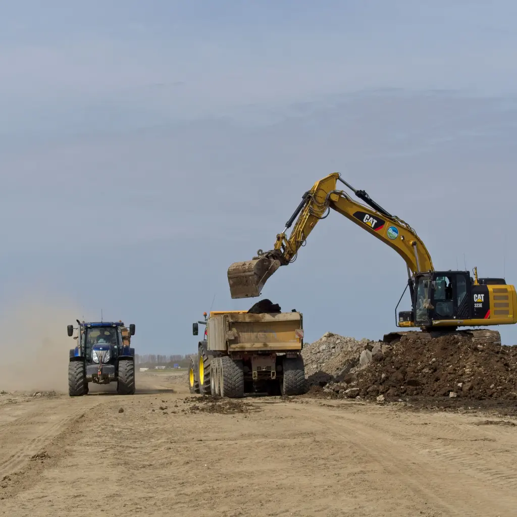 Excavator loads brown excavated earth into a dump truck on a dusty construction site.