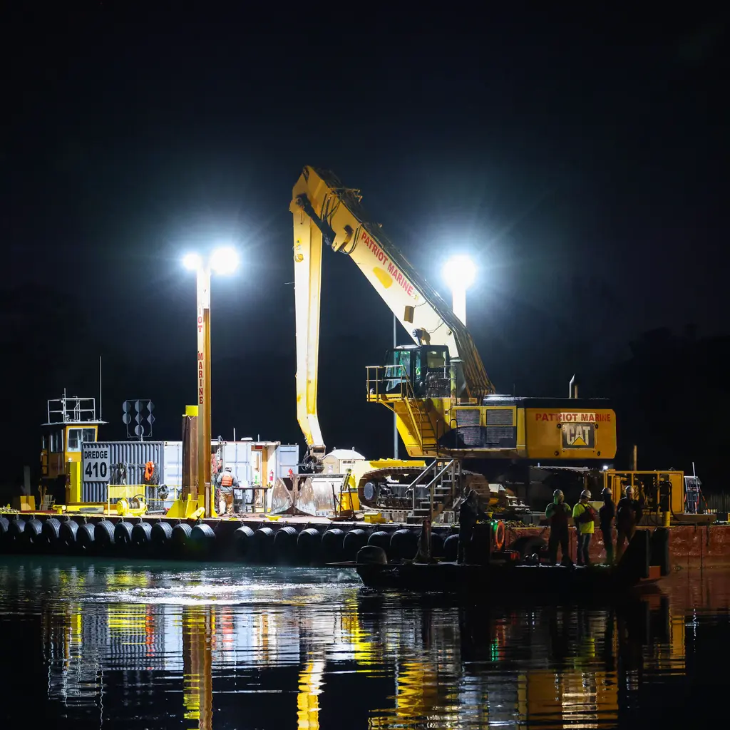 Floodlit dredging excavator works from a barge on dark marina water at night.