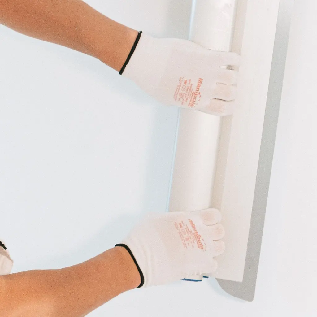 Gloved hands positioning a long straightedge against a smooth interior wall during drywall finishing.