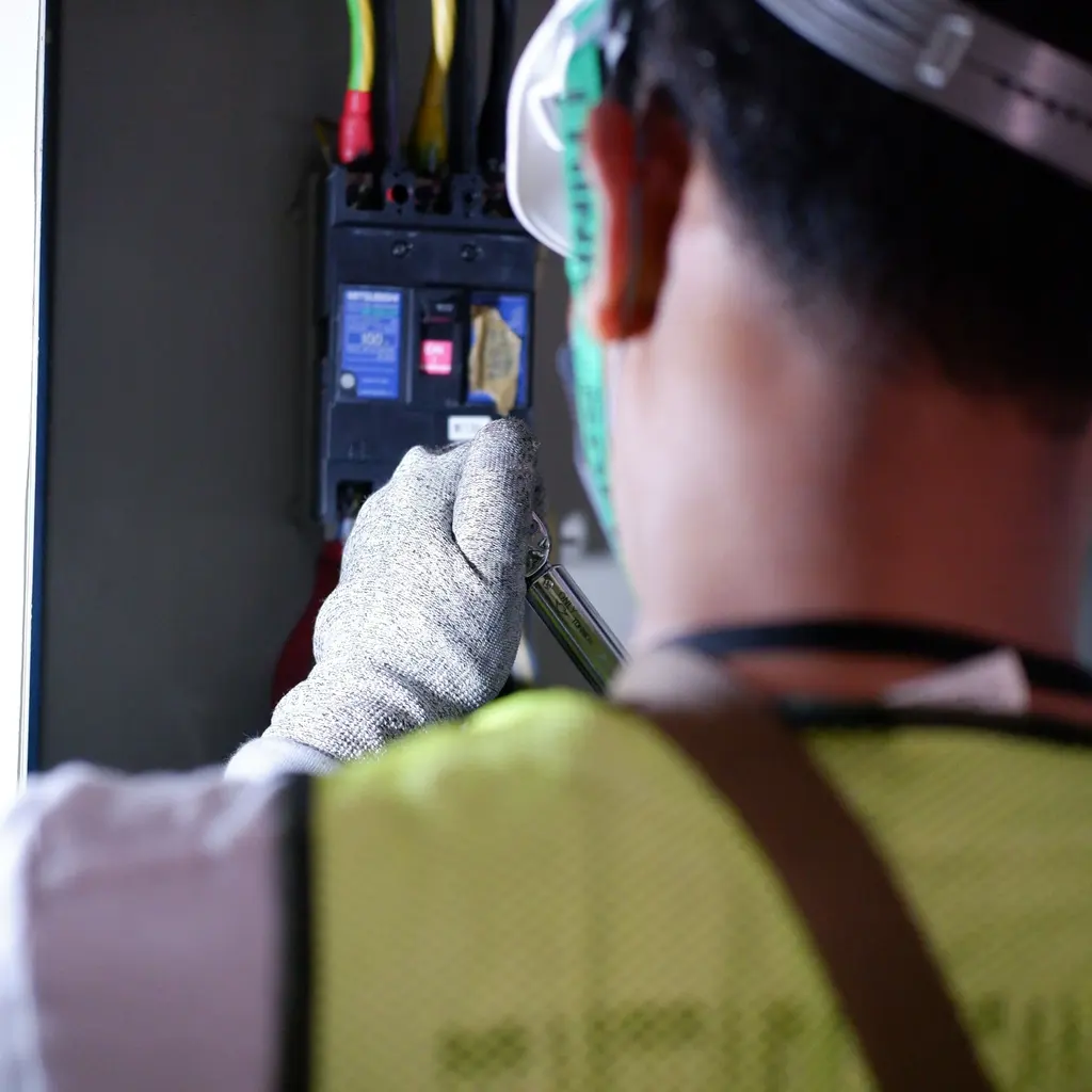A gloved electrical worker connecting wiring inside an open circuit breaker enclosure, viewed from behind.
