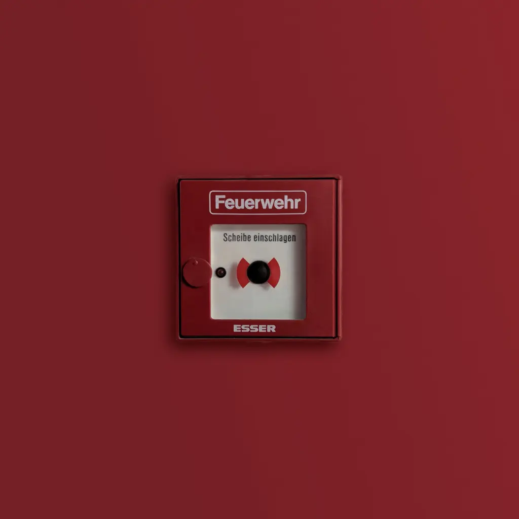 A red wall-mounted fire alarm call point set into a matching red panel, with its activation button visible.