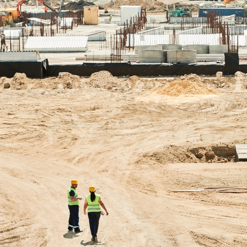 Two distant construction workers crossing a building site with foundations, reinforcing steel and earthworks behind them.