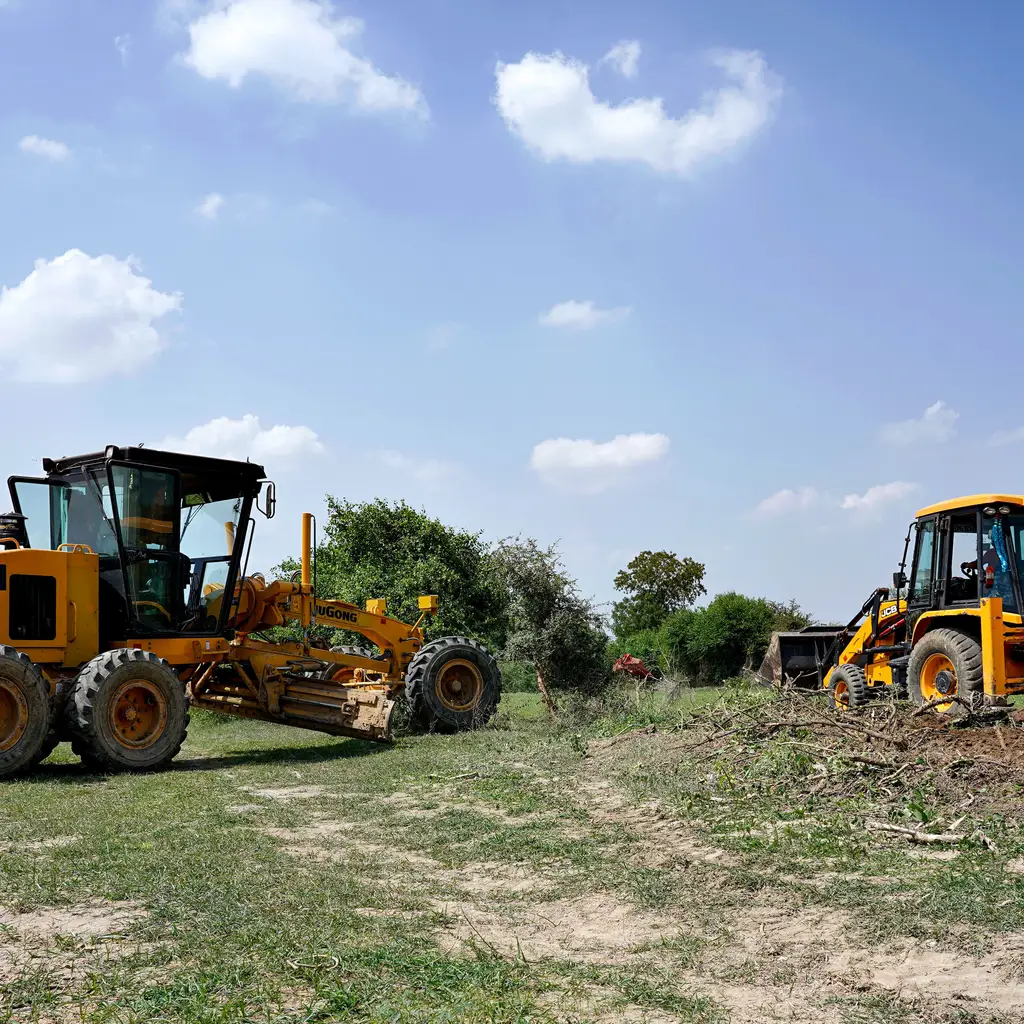 A yellow motor grader and a backhoe positioned on earth and grass at a land-development site.