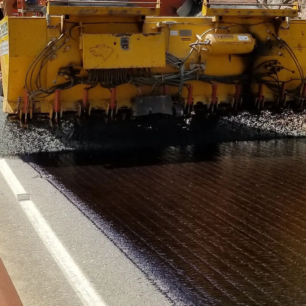 Road-surfacing equipment spreads dark aggregate beside a concrete curb.