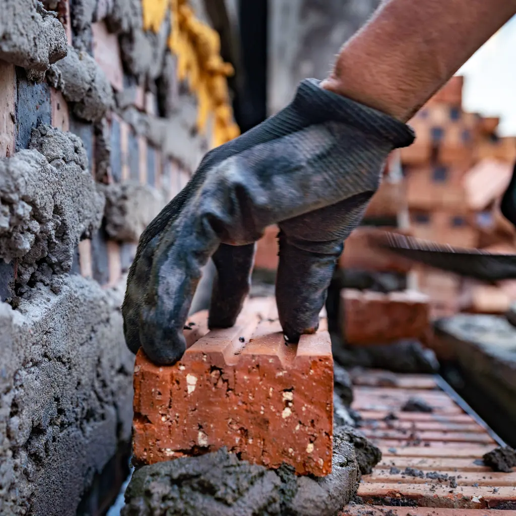 Gloved mason’s hand positioning a red brick along a mortar-covered wall course.