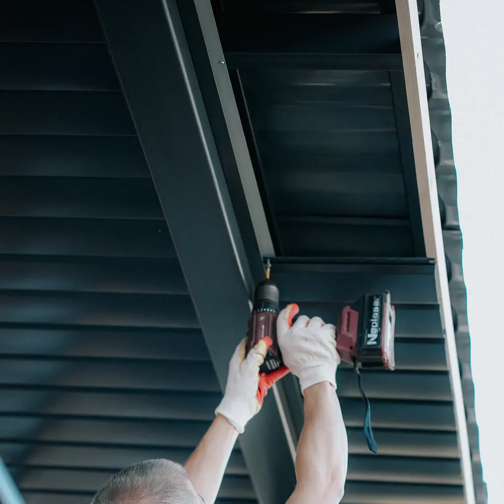 Gloved hands fastening dark metal roof panels from beneath a building overhang.