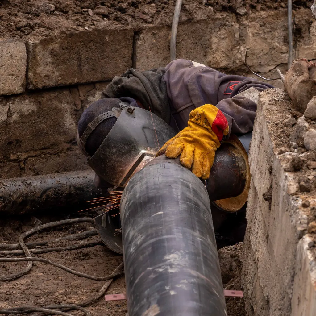 Mobile welder in a protective helmet working on a large steel pipe in an excavated trench.