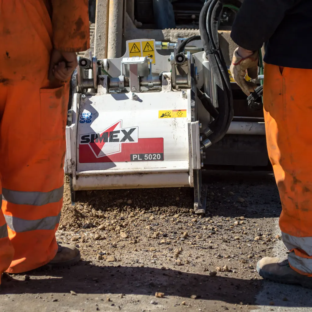 Road workers’ lower bodies beside asphalt-surface machinery and prepared road aggregate.