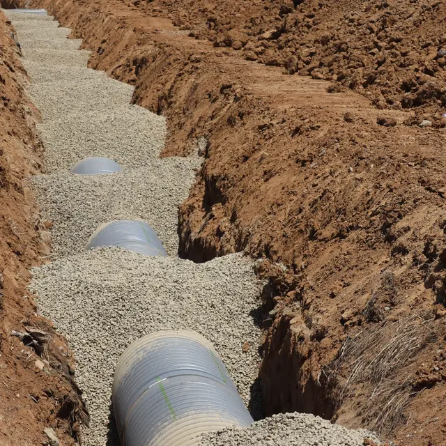 Drainage pipe sections aligned on gravel at the bottom of an excavated utility trench.