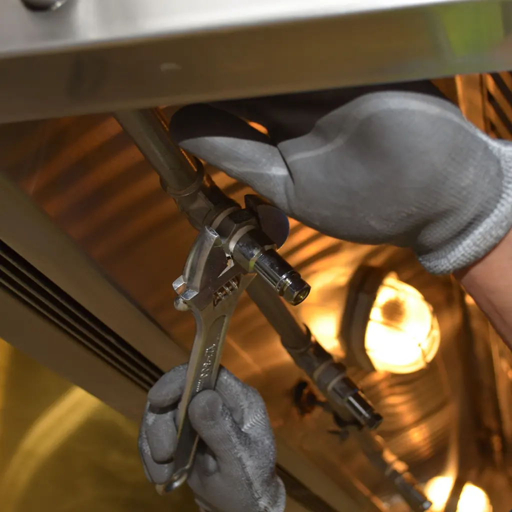 Gloved hands tightening an overhead pipe fitting with an adjustable wrench.