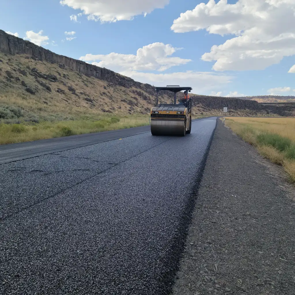 Road roller compacts fresh asphalt on a rural road bordered by dry hills.