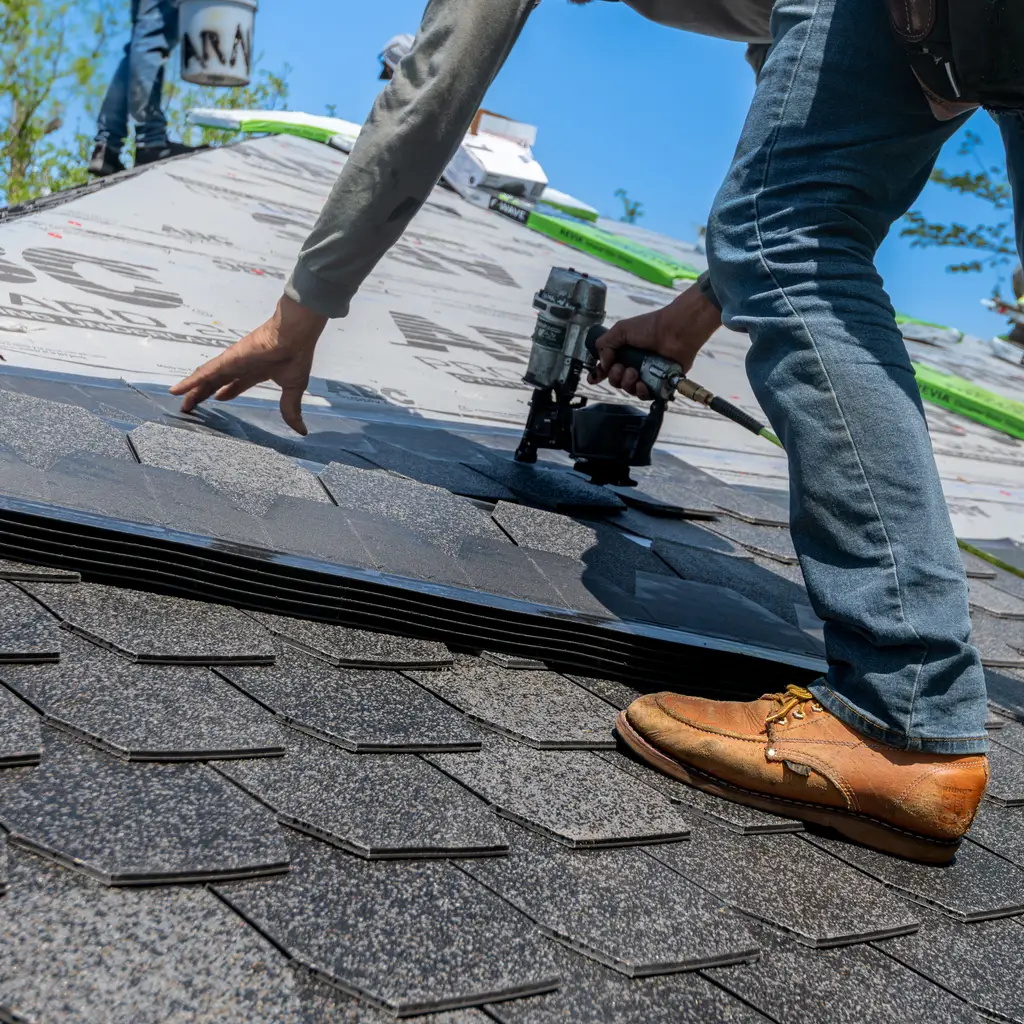 Roofer fastens asphalt shingles with a pneumatic nail gun on a sloping roof.