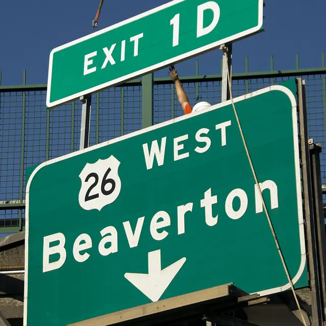 Large highway direction-sign panels hang from lifting cables while being raised into position beside a structure.