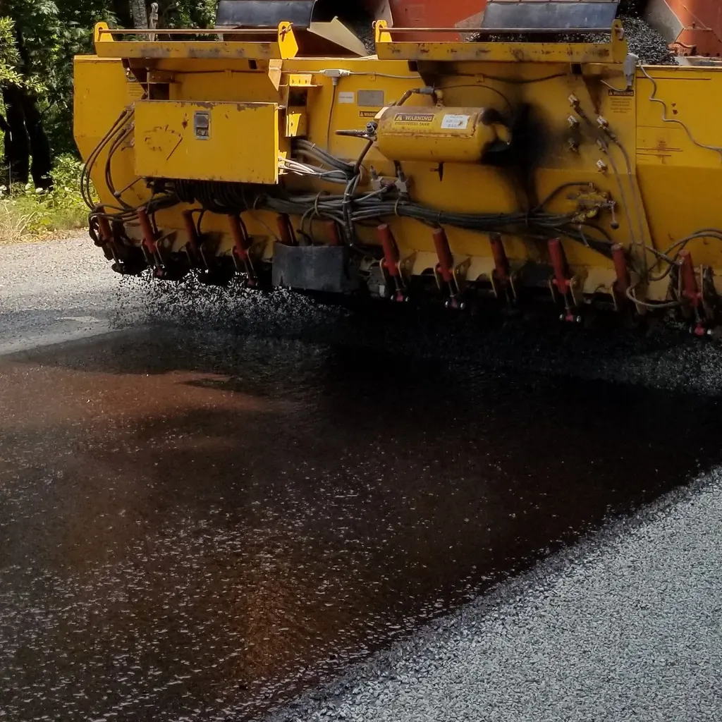 A road-surfacing machine spreads dark aggregate across a prepared road.