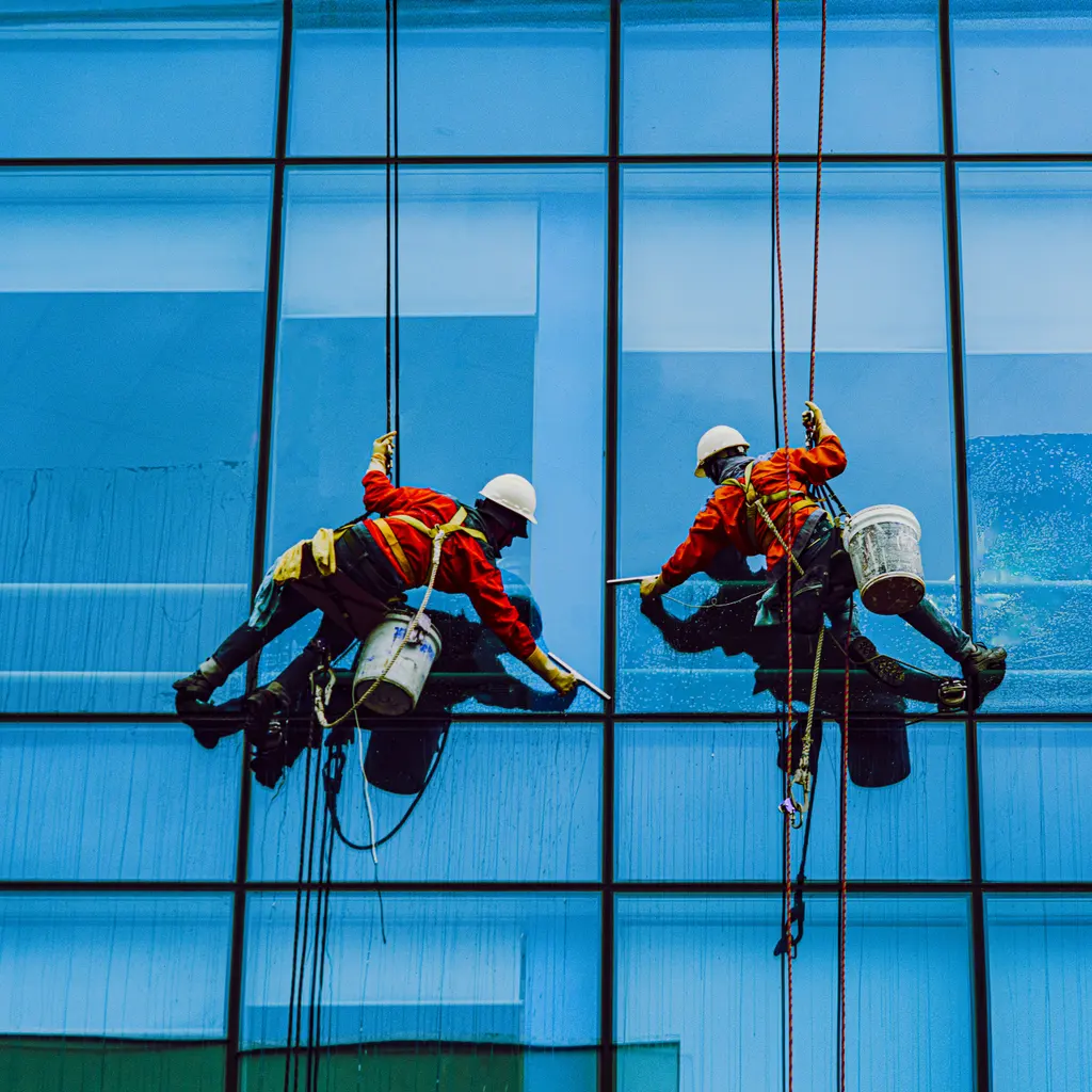 Two rope-access window cleaners wash a blue glass facade while suspended in safety harnesses.