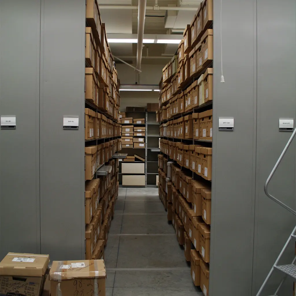 Archive boxes arranged on tall shelves along an aisle between metal storage cabinets.