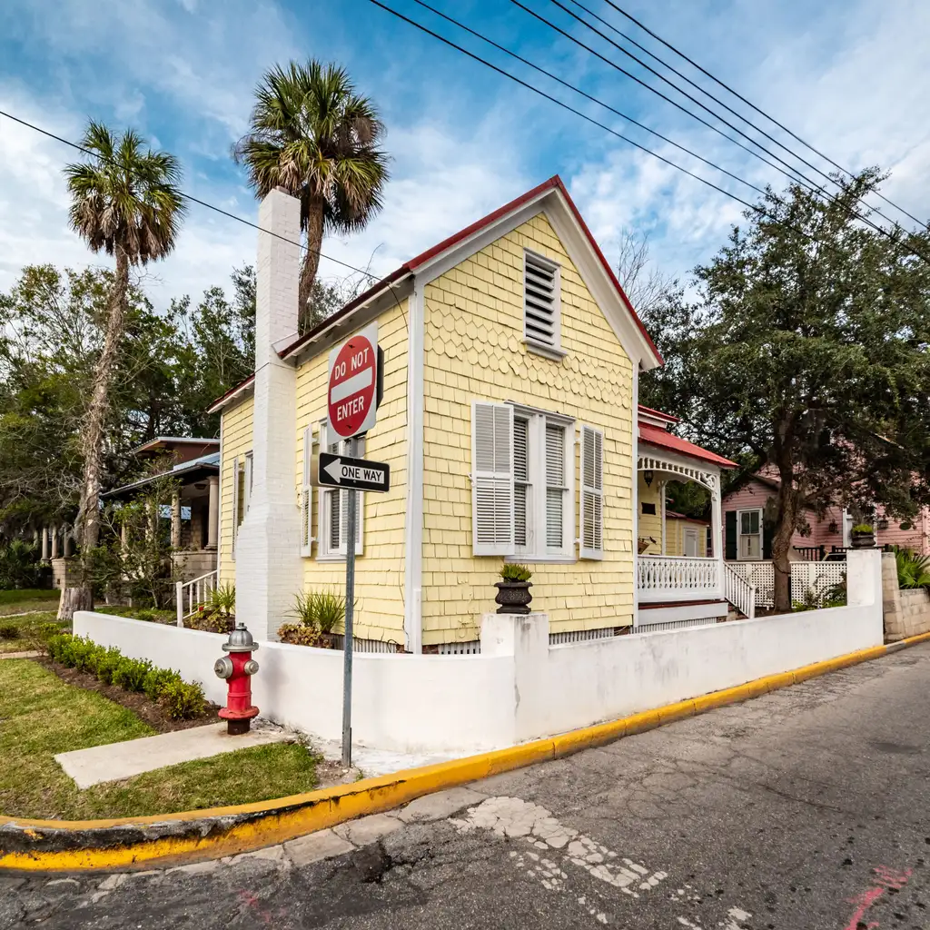Yellow house with white shutters and a front porch at a palm-lined street corner.