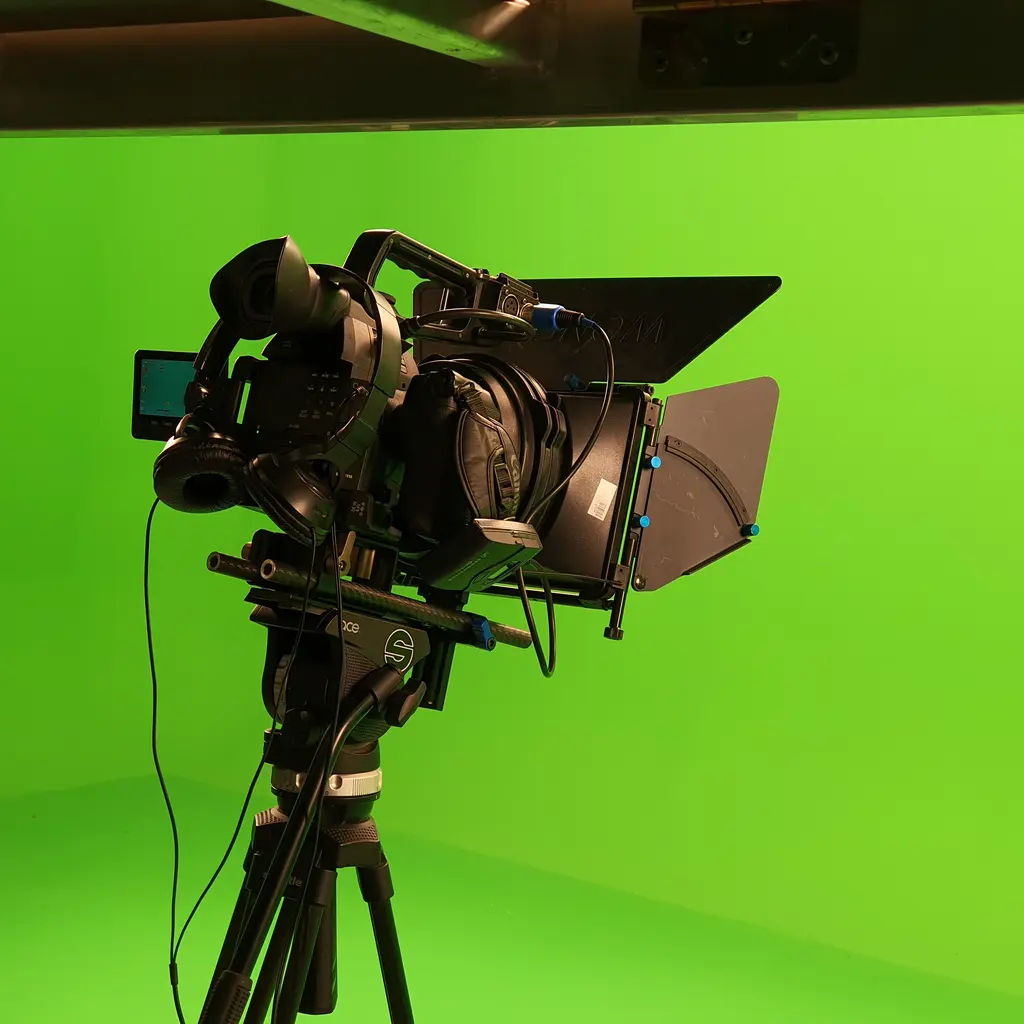 A video camera with a matte box and headphones stands on a tripod in a green-screen studio.