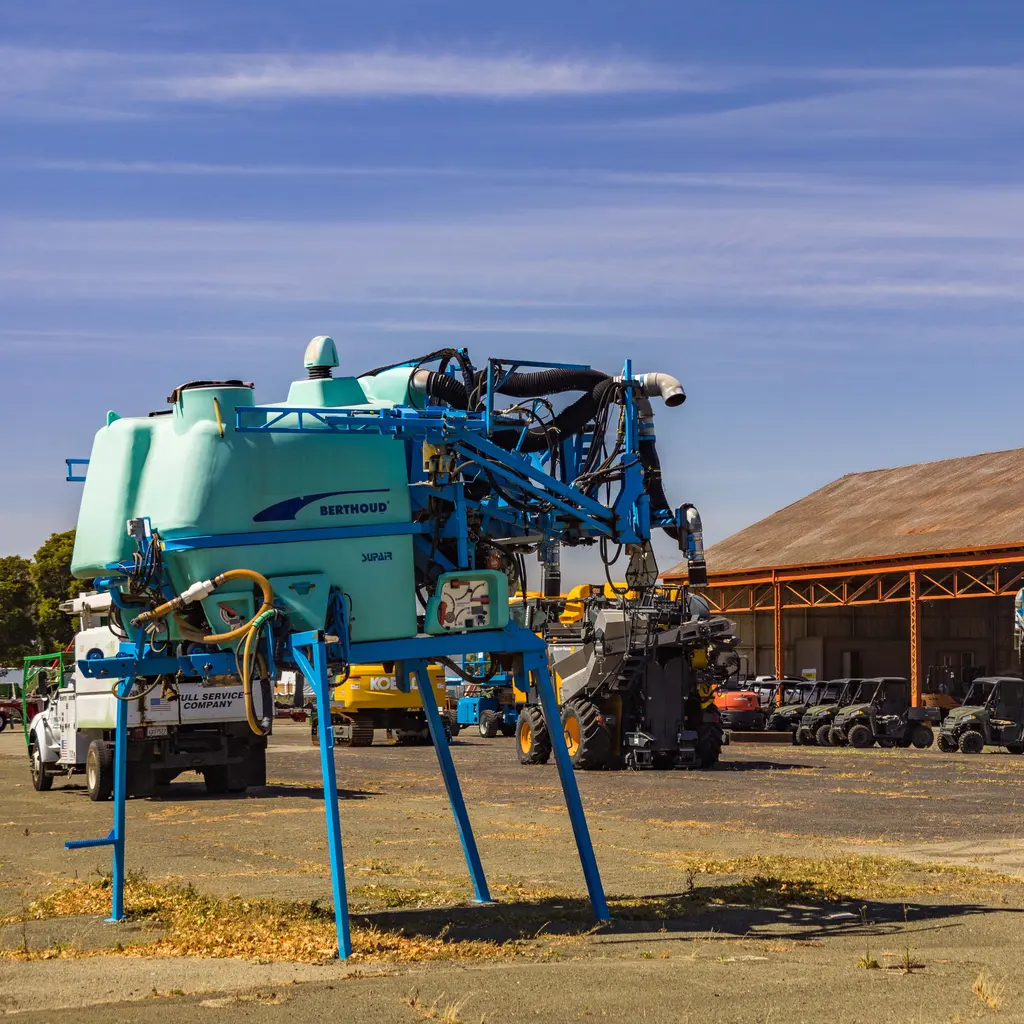Agricultural sprayer equipment with a large tank, hoses and folded booms in an equipment yard.