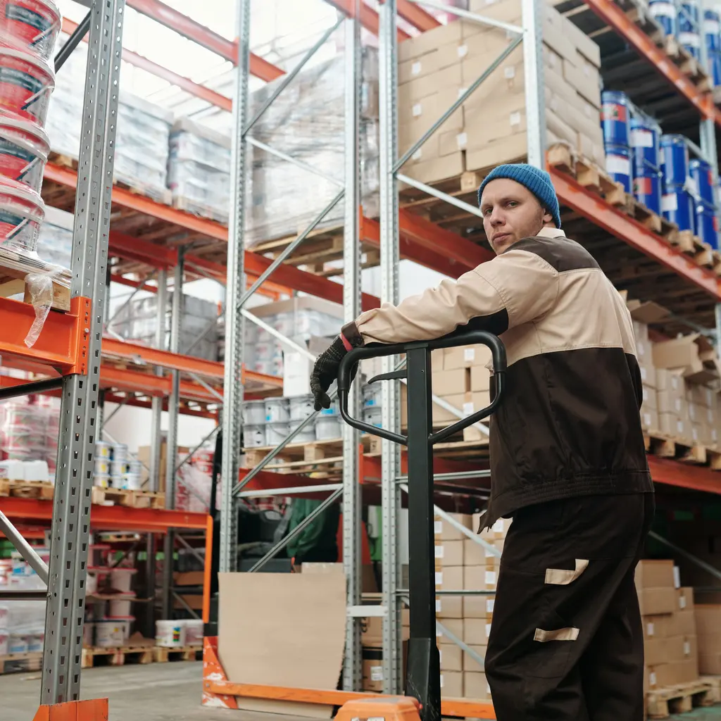 A warehouse worker moving a pallet jack beside tall racks of cartons and wrapped stock.