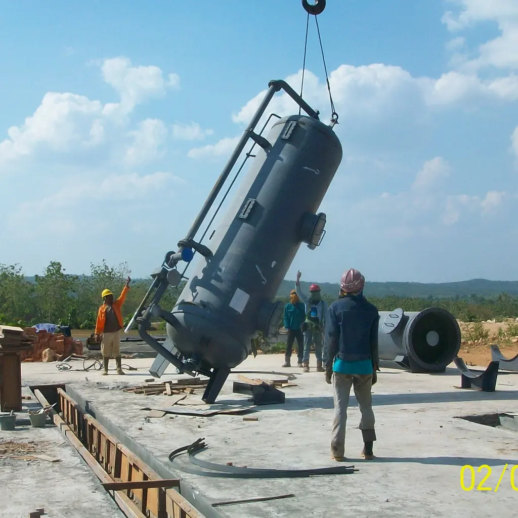 A vertical fired boiler being hoisted into position while workers guide it on a construction site.