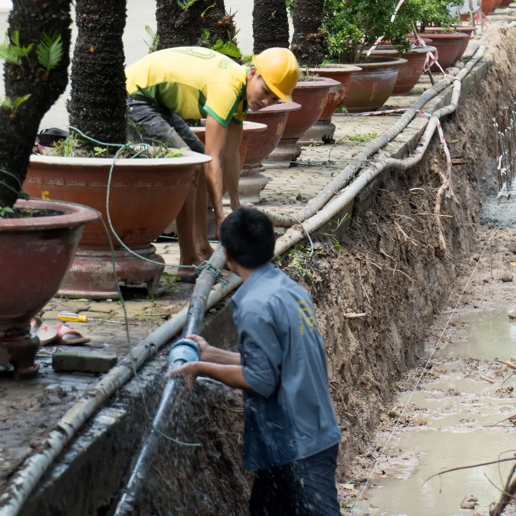 Workers fitting a water pipe along a narrow excavated trench beside a landscaped path.
