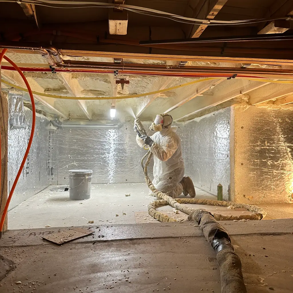A worker in protective clothing spraying foam insulation onto the underside of a crawlspace ceiling.