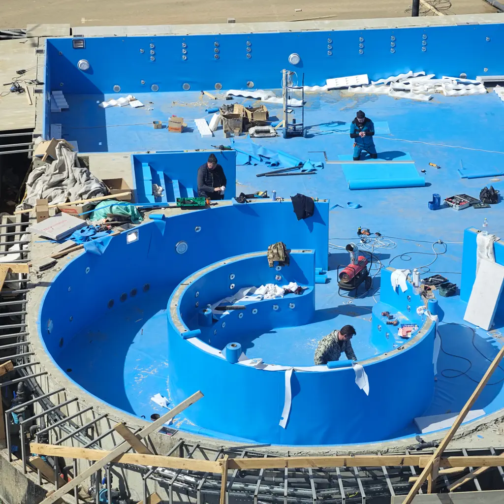 Workers fitting the blue waterproofing and built-in features of an unfinished swimming pool.