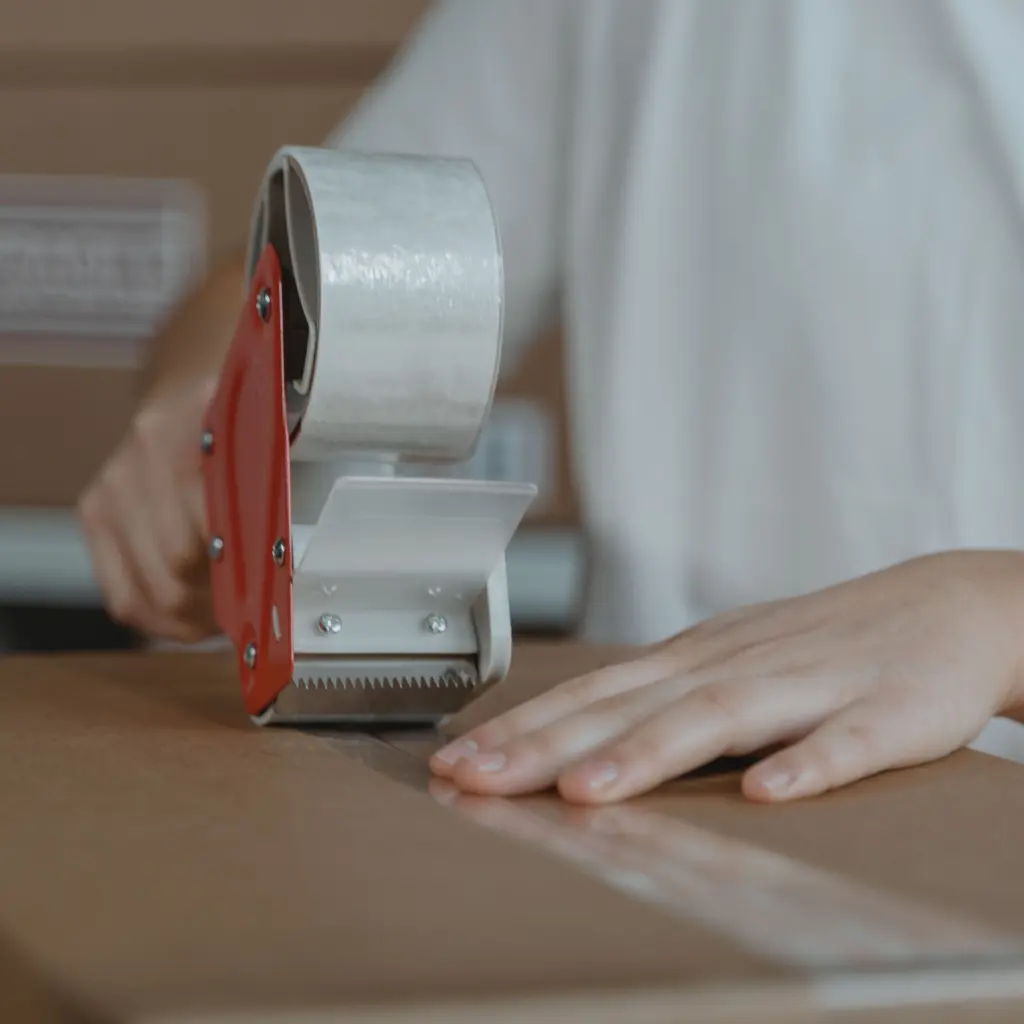 Hands sealing a cardboard shipping carton with clear packing tape.