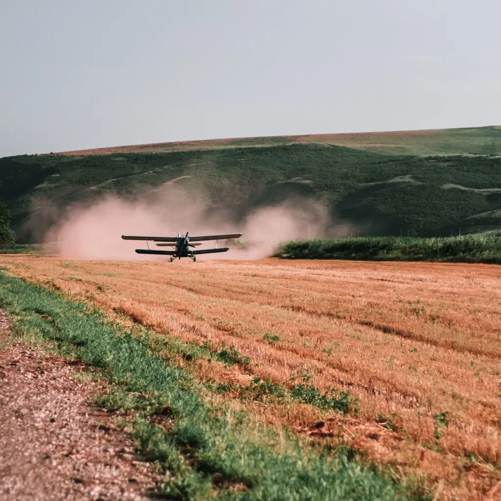 A crop-dusting aircraft flying low over a field, with a plume trailing behind it.