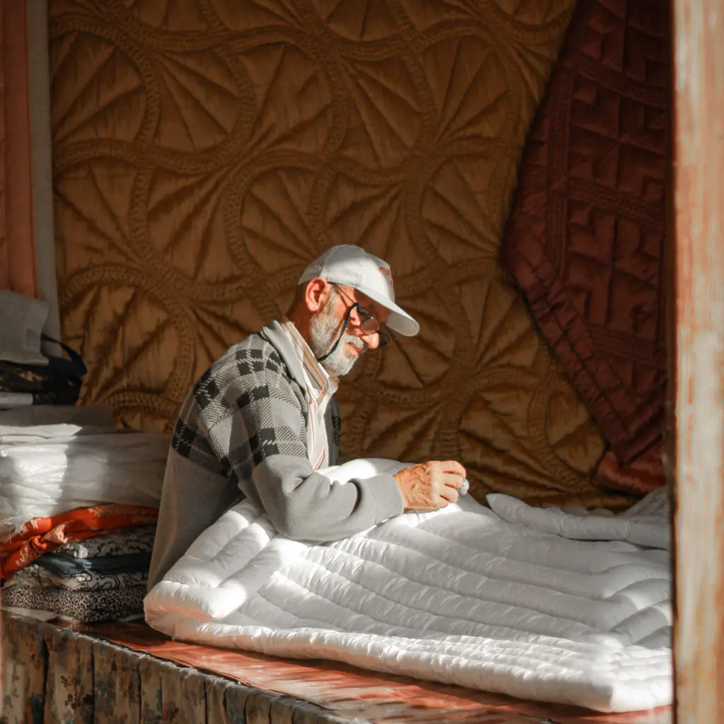 A person stitching a thick white quilt with other quilted textiles hanging behind the workbench.