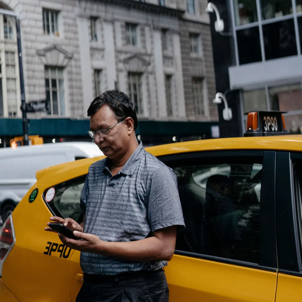 A taxi driver using a smartphone beside a yellow cab on a city street.