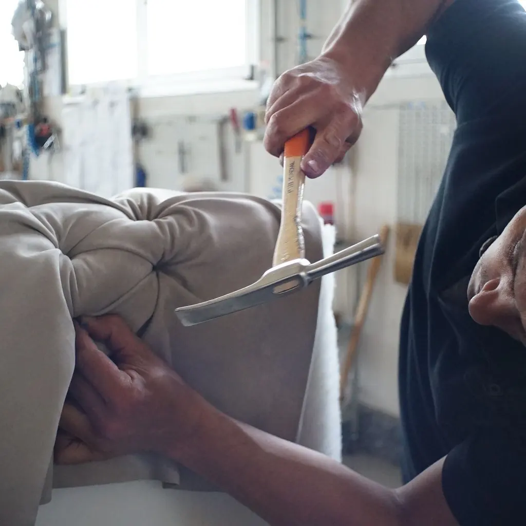 An upholsterer fastening fabric along the edge of a tufted sofa with a small hammer.