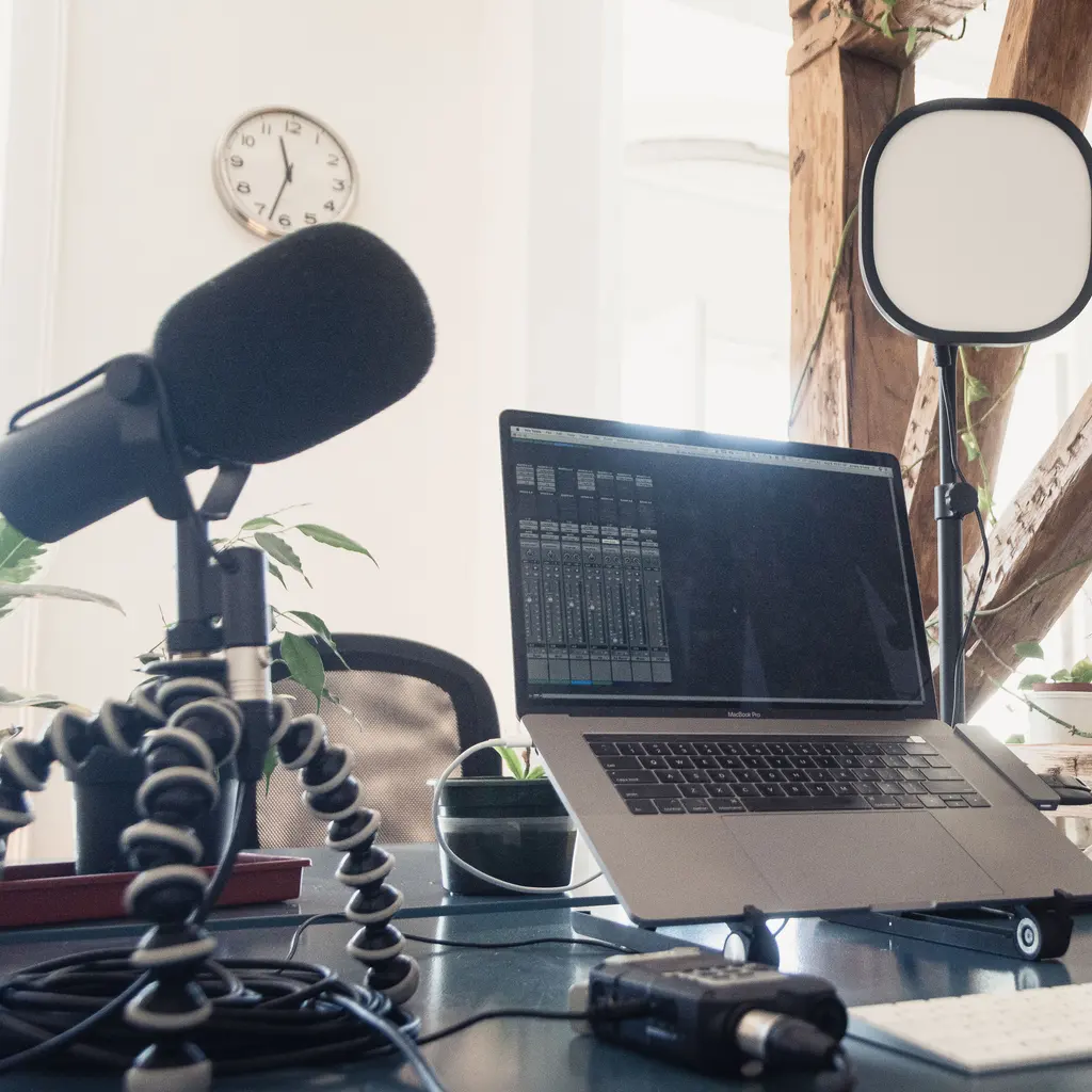 A tabletop microphone stands beside a laptop displaying audio-mixer controls in a podcasting workspace.