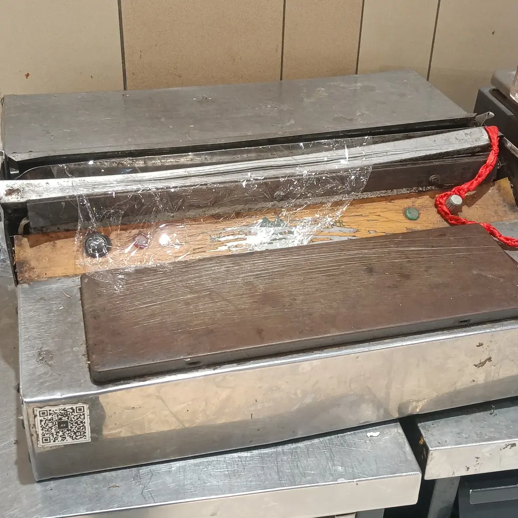 A stainless steel plastic-film wrapping machine with a roll of clear film above the work surface.